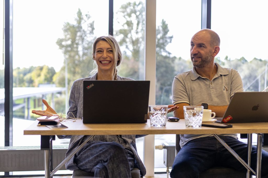 Two people sit in a conference room, with large windows behind them. On the tables in front of them are computers and drinking glasses. A woman is laughing and moving her hands; a man is listening and smiling.