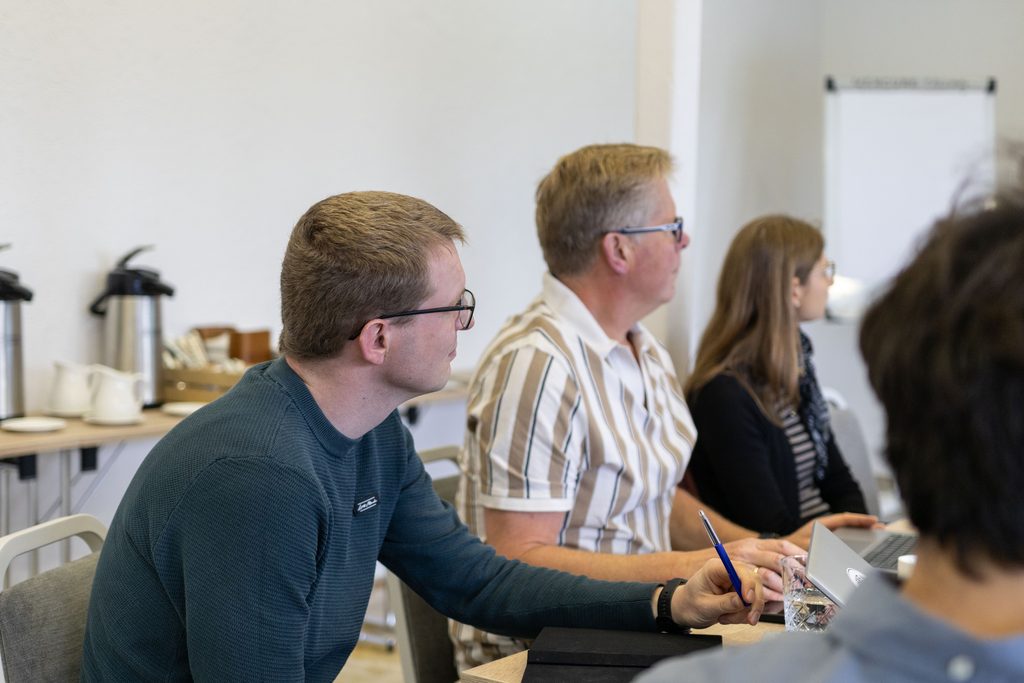 Three people sit in a conference room around a table and listen to a presentation. Behind them is a table with coffee.