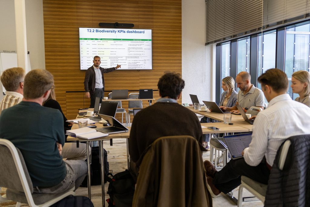 People are sitting in a conference room and listening to a man present in front of the room. The man is presenting a slide "Biodiversity KPIs and dashboard" on the screen.