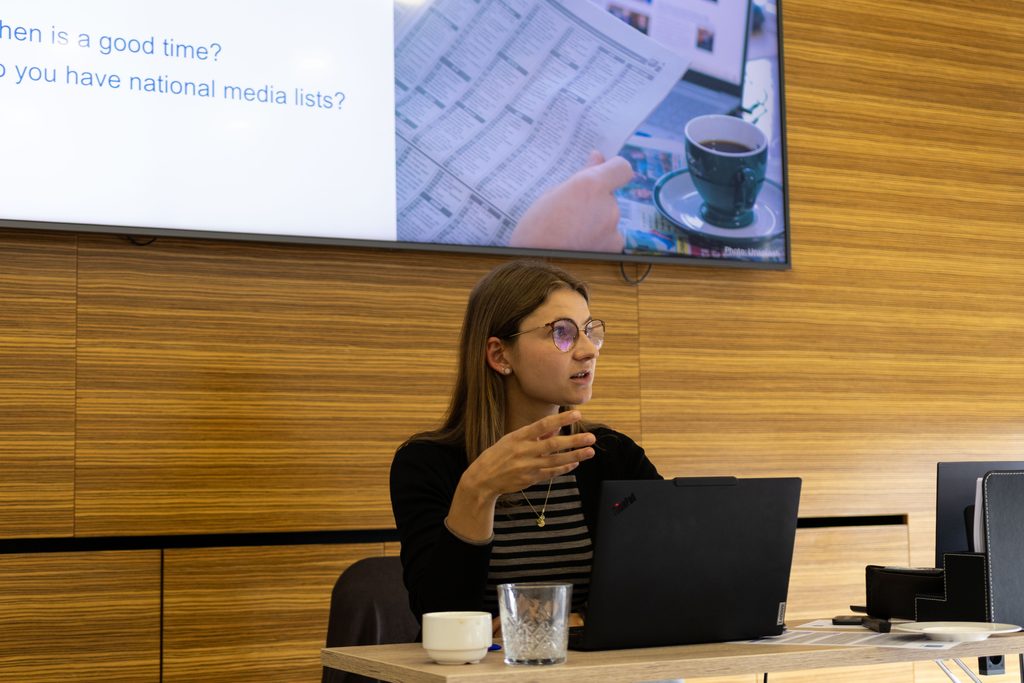 A woman is sitting behind a table and presenting something. In front of her is a laptop, coffee cup and a glass of water. Behind her is a presentation featuring an image of a person reading a newspaper.